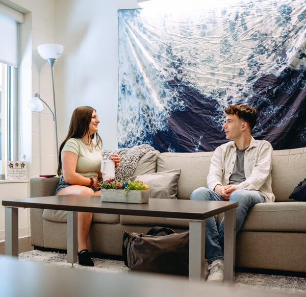 Two students talk on a couch in a common living area with a large, blue tapestry on the wall behind them